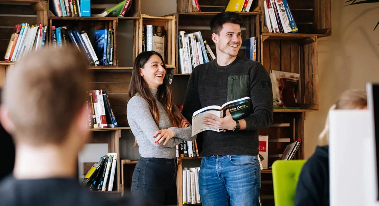 2 Personen stehen vor einem Bücherregal und lachen, die rechte Person hat ein Buch in der Hand.